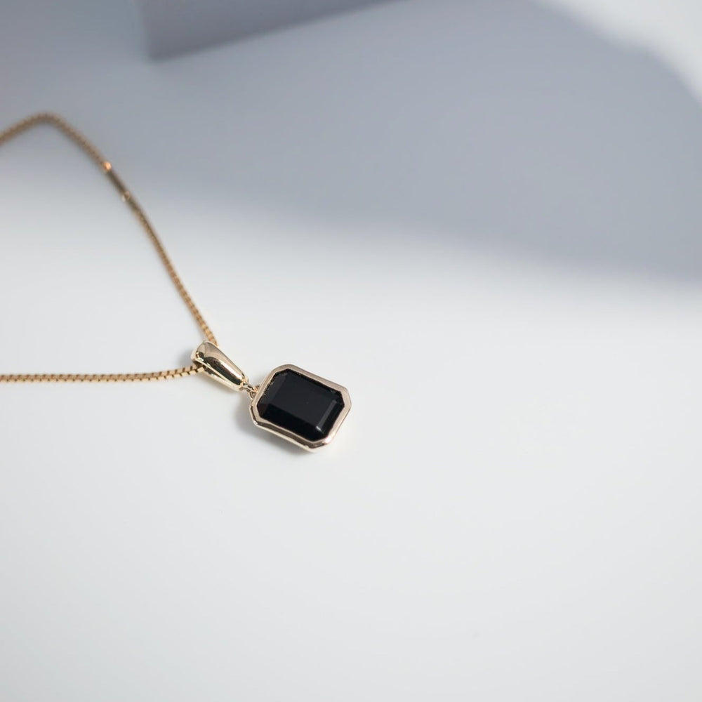 
                      
                        A delicate Estelle Necklace by Pear rests on a white surface, its gold chain highlighting the elegance. The pendant showcases a rectangular black onyx stone encased in a solid gold bezel. Soft lighting casts gentle shadows, enhancing the elegant and minimalist atmosphere of the image.
                      
                    