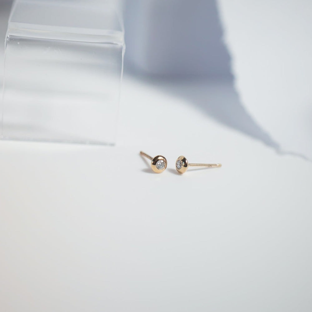 A close-up of a pair of small, minimalist Juliette Bezel Earrings from Pear, featuring a 14K gold bezel and a single round gemstone in the center of each. The diamond studs are placed on a white surface with soft lighting, casting delicate shadows.