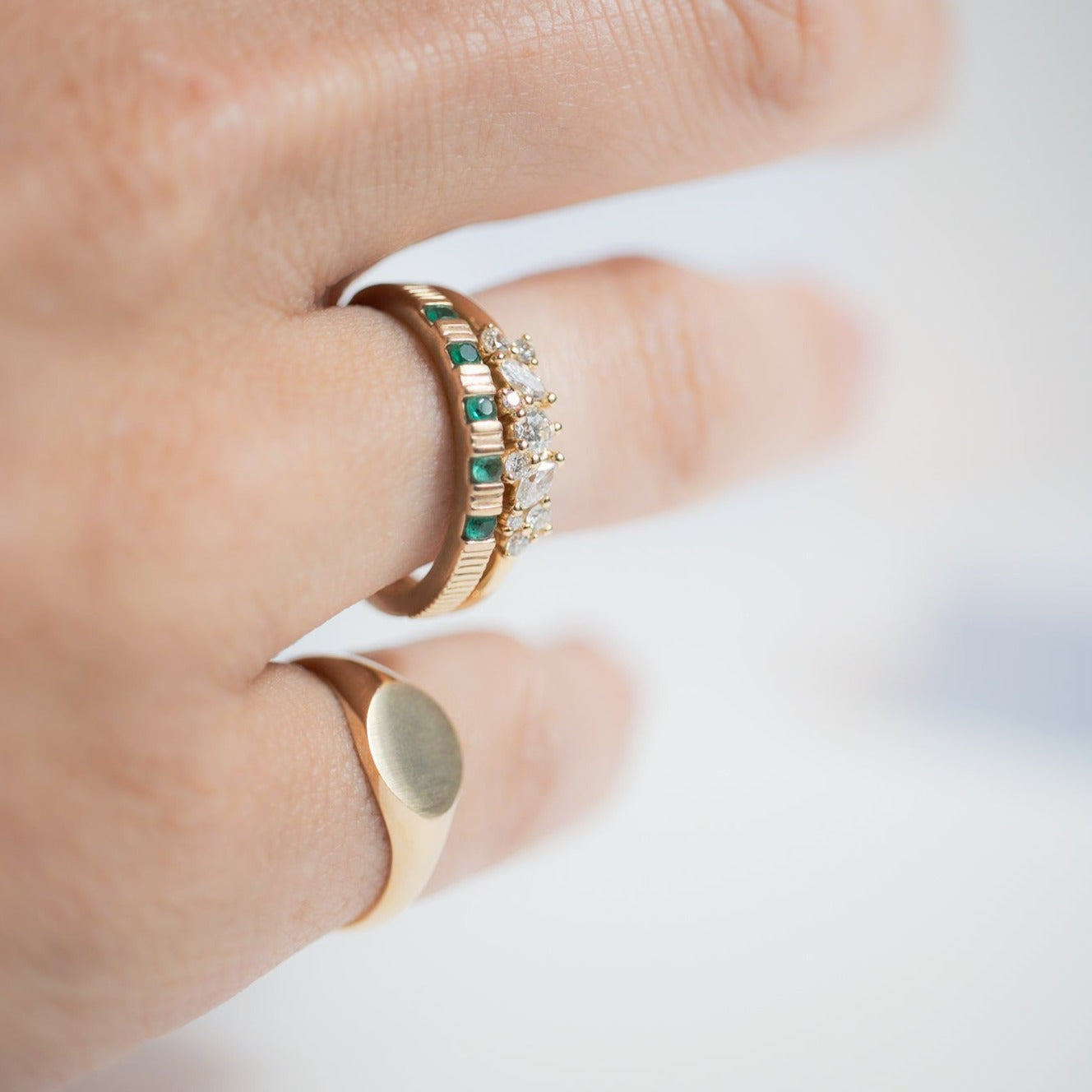 A close-up of a hand adorned with three elegant gold rings from Pear, each distinct in its charm. One ring, the Gemstone Ribbed Ring, showcases small emerald gemstones and diamonds. Another ring features a smooth, plain oval design. The third ring is a simple band embellished with a subtle ribbing design. The focus remains on the rings, highlighting their intricate details.
