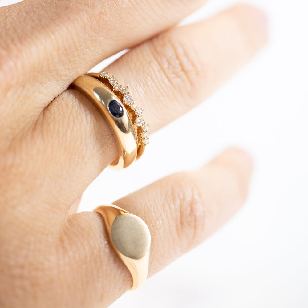 
                      
                        A close-up of a light-colored hand wearing three golden rings. The middle finger is adorned with two rings: one thick gold band featuring a black stone and another thin band embedded with small white stones. The ring finger displays the Petite Round Signet Ring by Pear, an engraved, thick gold signet ring.
                      
                    