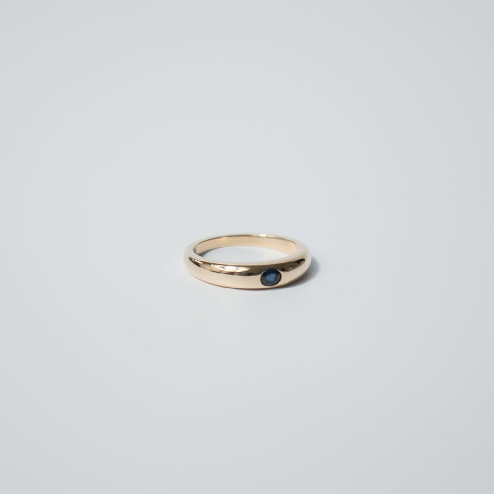 
                      
                        The Sapphire Dome Ring by Pear features a smooth, polished band crafted in 14K gold, set with a small blue sapphire in the center, placed against a plain light gray background.
                      
                    