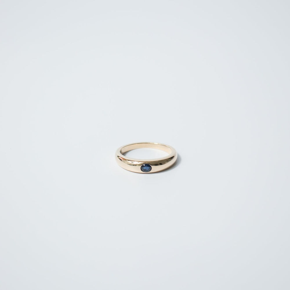 
                      
                        The Pear Sapphire Dome Ring, featuring a single round dark blue sapphire set in the center, lies on a smooth, light gray surface. Crafted from 14K gold, the ring boasts a simple and elegant design.
                      
                    