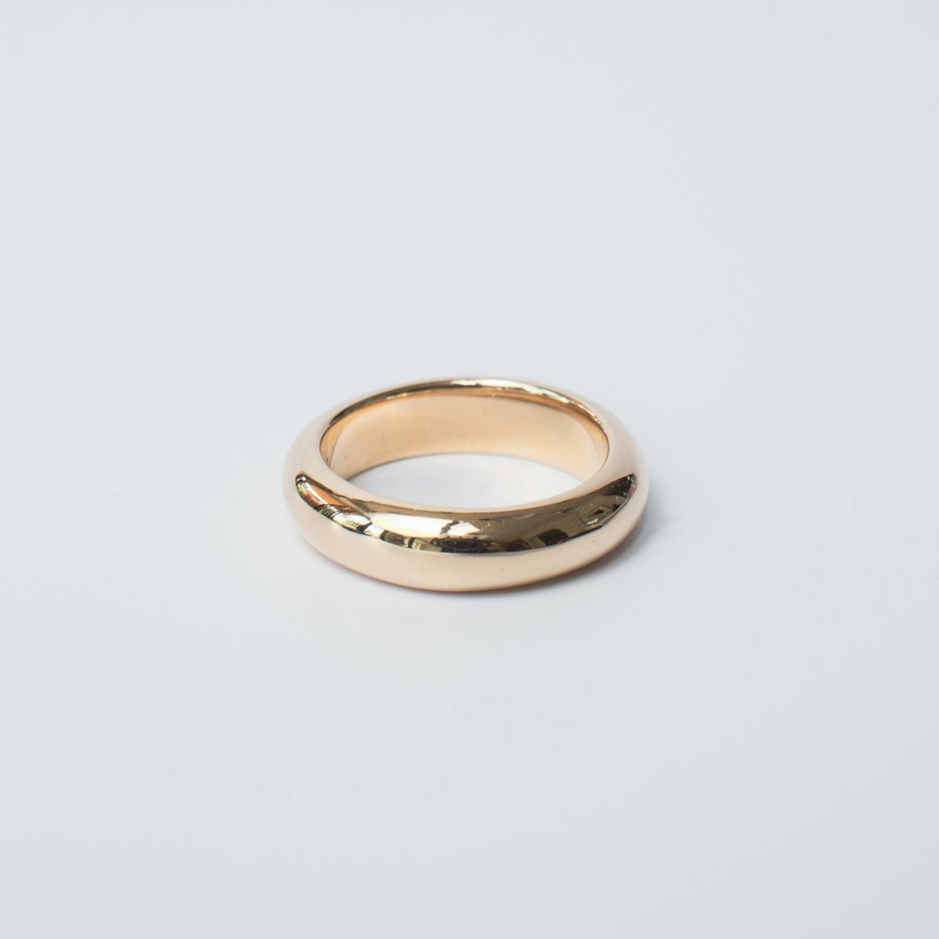 A polished, solid gold band known as the "Heavy Band" by Pear, featuring a smooth, rounded design, is positioned on a plain, light background. The ring boasts a reflective surface and its simple, elegant appearance suggests it could be a classic wedding band or an exquisite piece of everyday jewelry.