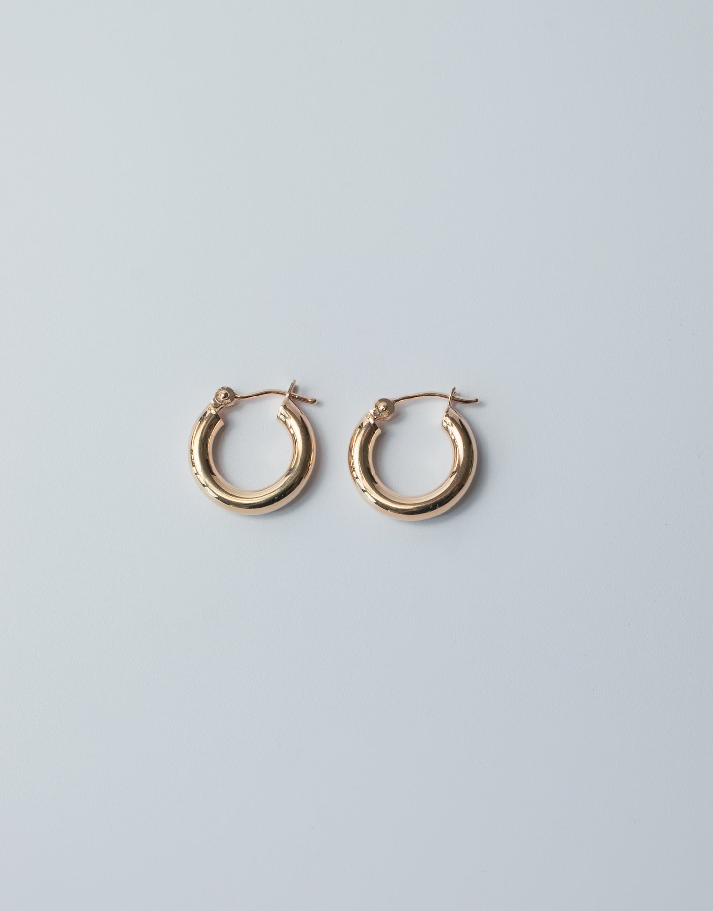 The Mini Thick Hoops by Pear, a pair of small 14K gold hoop earrings, are elegantly displayed on a light gray background. These mini hoops feature a plain, smooth, polished surface and latch backs for secure closure. Their simplicity and shine make them perfect for everyday wear, standing out beautifully against the neutral backdrop.