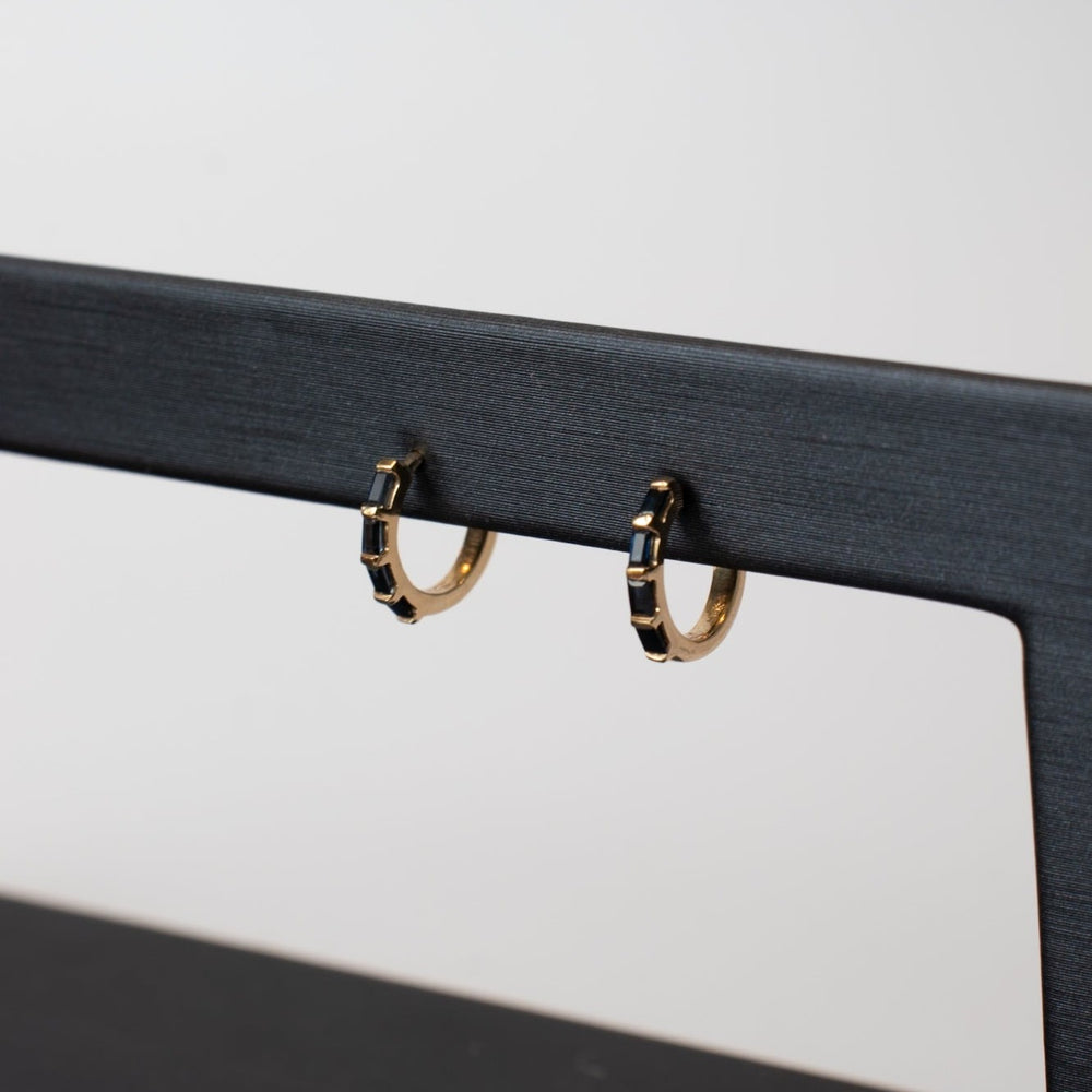 
                      
                        A pair of Sapphire Huggies from Pear featuring gold hoops with black geometric designs, elegantly draped on a sleek, black jewelry display stand. The plain background accentuates the earrings, making them an elegant everyday staple.
                      
                    