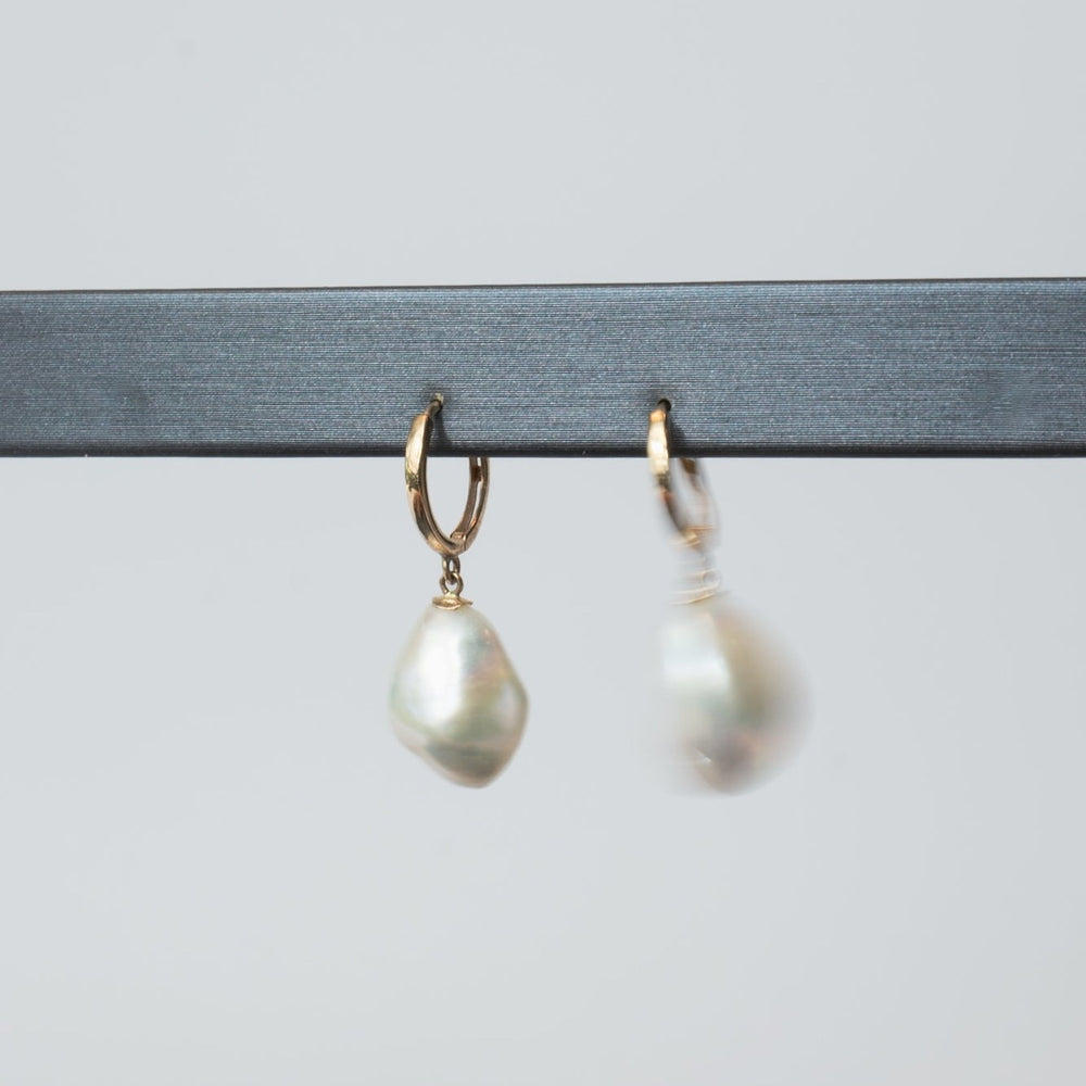 
                      
                        A close-up image of two Pearl Accented Hoops by Pear showcases each gold earring adorned with a single irregular, pear-shaped freshwater Keshi pearl. One earring is in sharp focus while the other appears slightly blurred against a plain, light gray background. These luxurious jewelry pieces hang elegantly from a black bar.
                      
                    