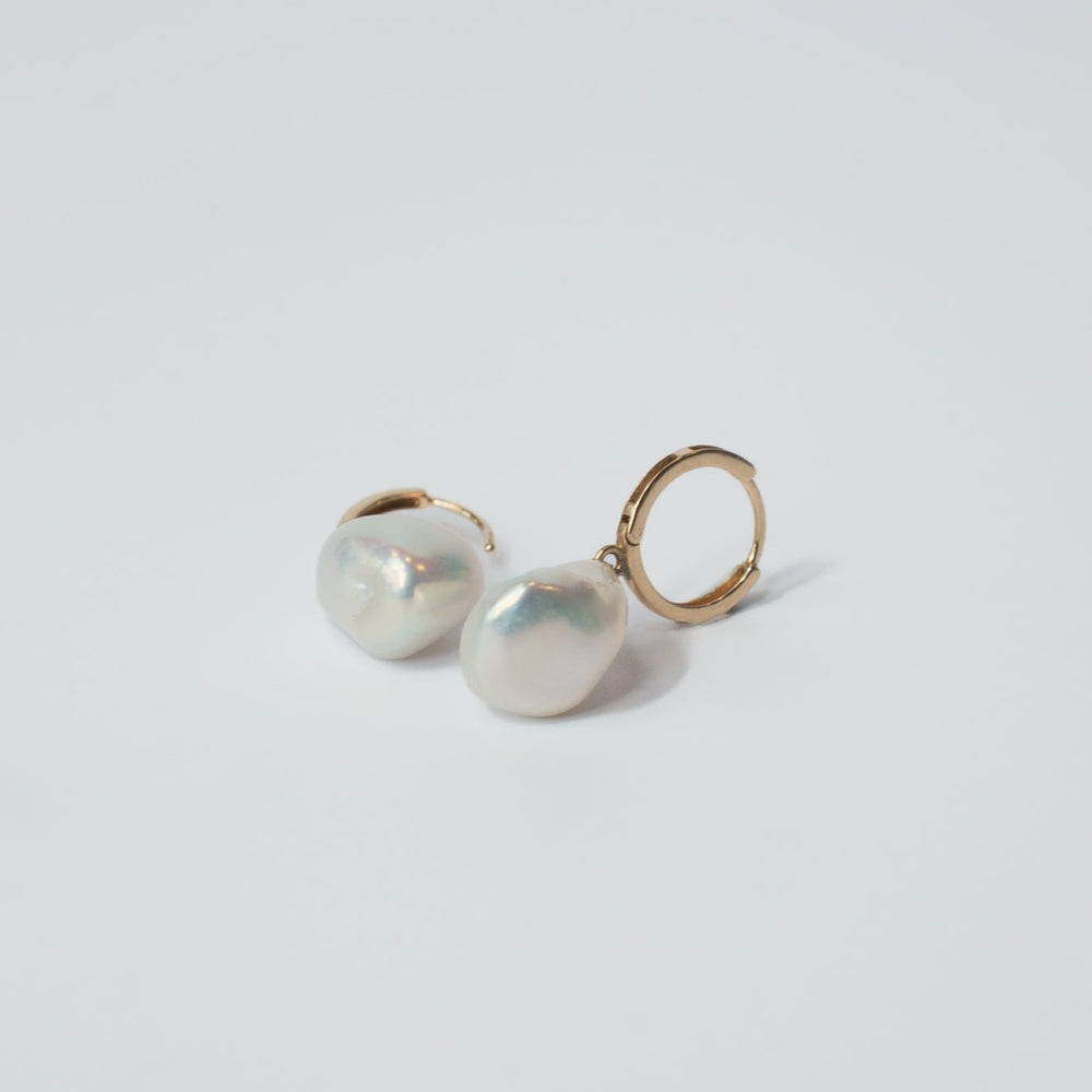 
                      
                        The Pearl Accented Hoops by Pear are a pair of 14K gold earrings, each featuring a large, irregularly-shaped freshwater Keshi pearl. Placed on a smooth, white surface with minimal shadows, these earrings elegantly showcase their simple design, making them a perfect addition to any luxury jewelry collection.
                      
                    