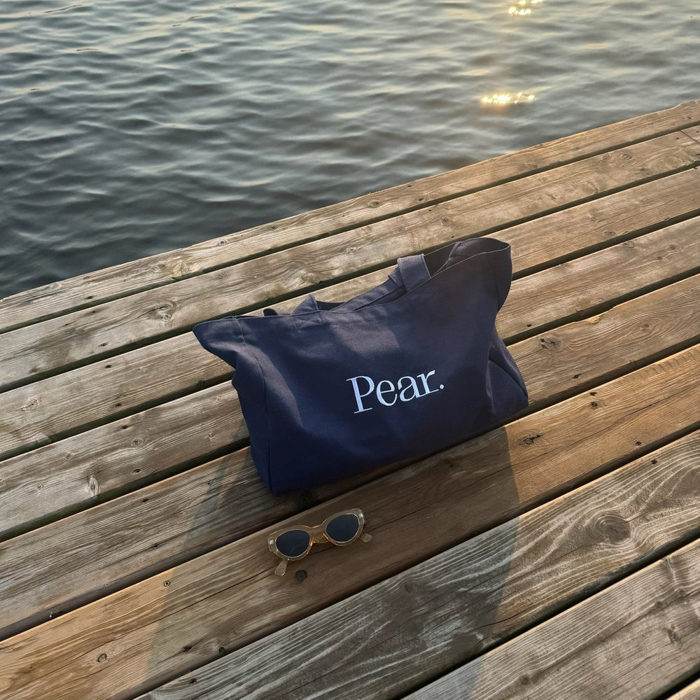 A navy blue Everyday Tote Bag from Pear. sits on a wooden dock near the water. A pair of sunglasses rests on the dock in front of the heavier weight, cotton canvas tote bag. The water reflects sunlight, creating a tranquil atmosphere.