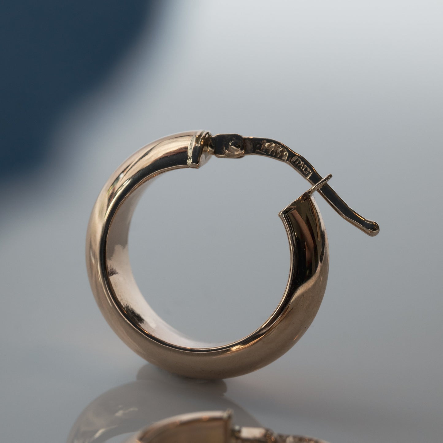 Close-up of a single Pear Solid Chunky Hoop earring with a smooth, polished surface. The solid half hoop is circular and features a hinged clasp for easy wearing. The background is blurred, directing all attention to the elegant lightweight earring from Pear.