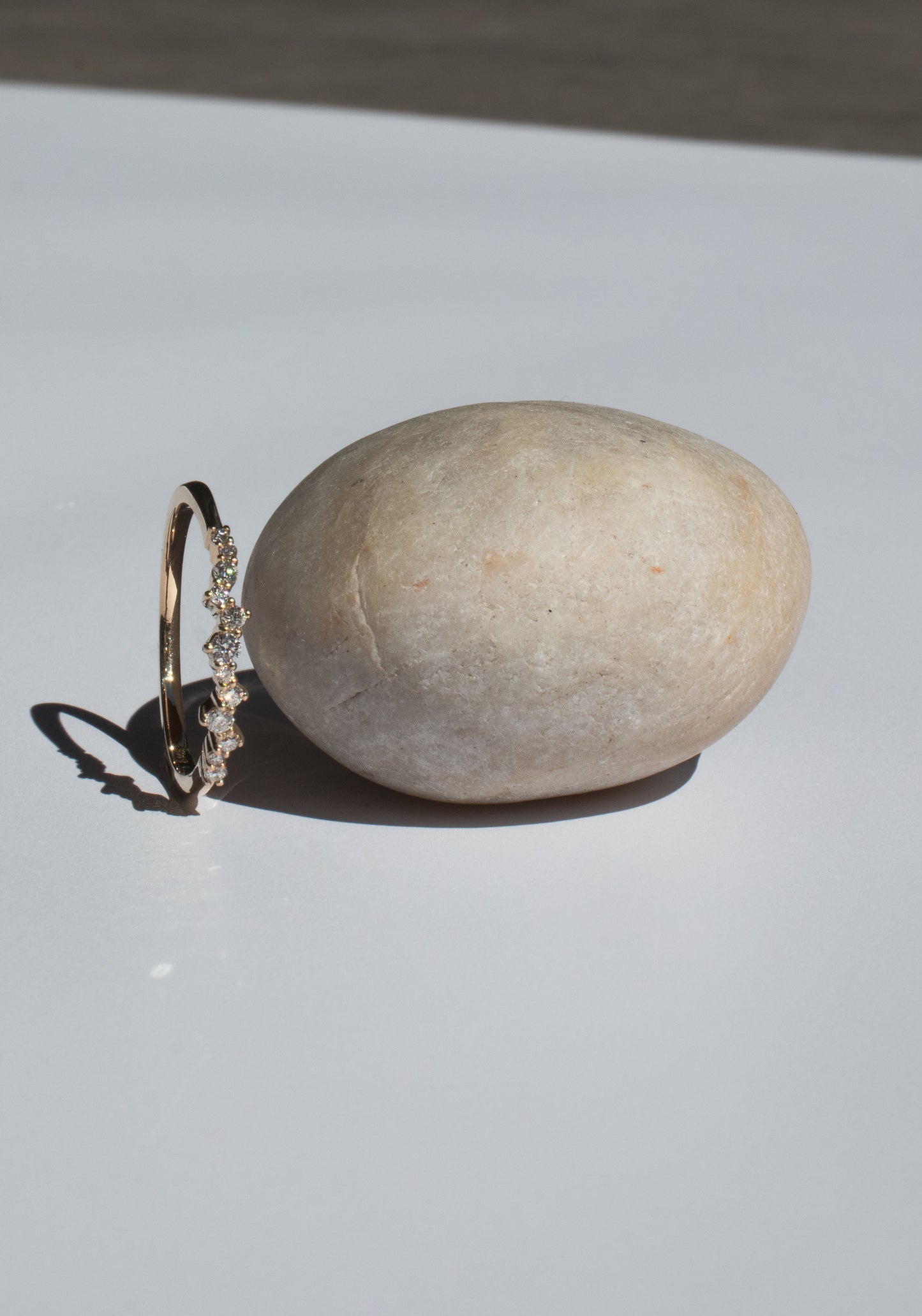 A delicate gold ring with small diamonds rests against a smooth, oval-shaped stone on a white surface. The lighting casts soft shadows, highlighting the ring's sparkle and the stone's texture.