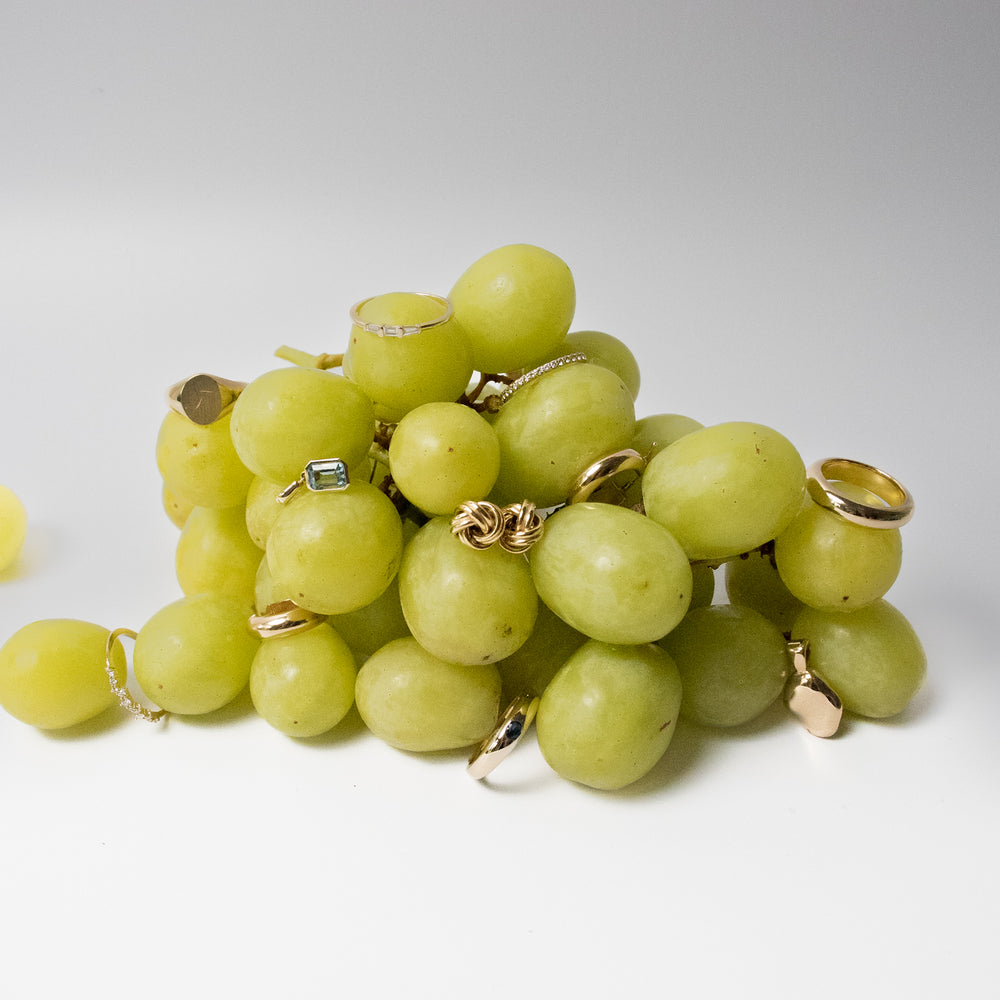 A cluster of green grapes is adorned with various gold rings, some with gemstones. One grape to the left is not decorated. The background is white and minimalistic, highlighting the unusual combination of grapes and jewelry.