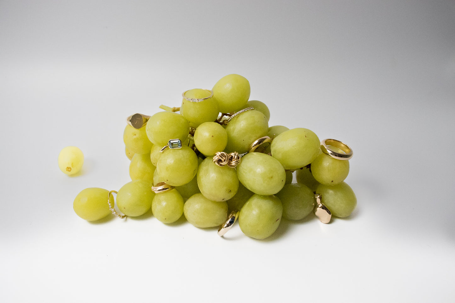 A cluster of green grapes is adorned with various gold rings, some with gemstones. One grape to the left is not decorated. The background is white and minimalistic, highlighting the unusual combination of grapes and jewelry.