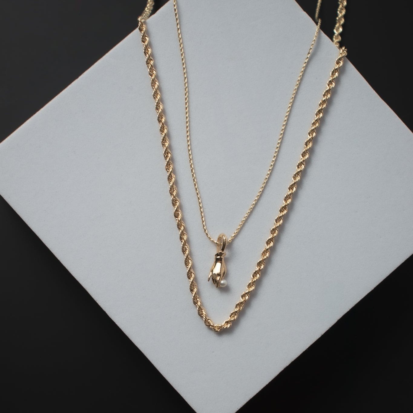 Two 14K yellow gold necklaces are displayed on a square white surface against a dark background. One necklace features a delicate boxing glove pendant, while the other is the "Round Rope Chain" by Pear., with a design reminiscent of a thicker, rope-like 2mm wheat chain.