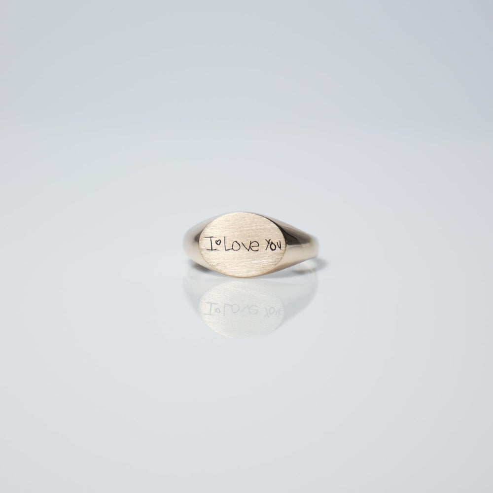 The Handwritten Oval Signet Ring by Pear., crafted in silver and featuring a horizontal oval design, sits elegantly on a smooth, reflective surface against a light gray background. Customize this ring with a laser-engraved handwritten message for that extra special touch.