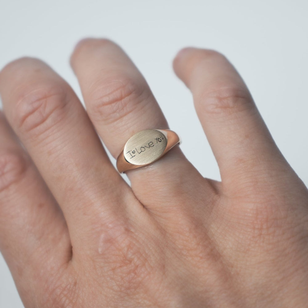 
                      
                        A close-up features a hand wearing the Handwritten Oval Signet Ring by Pear., showcasing the "Love You" engraving on its horizontal oval surface. This laser-engraved message adds a personal touch to the smooth ring design. The fingers are slightly bent, set against a softly blurred, light-colored background.
                      
                    