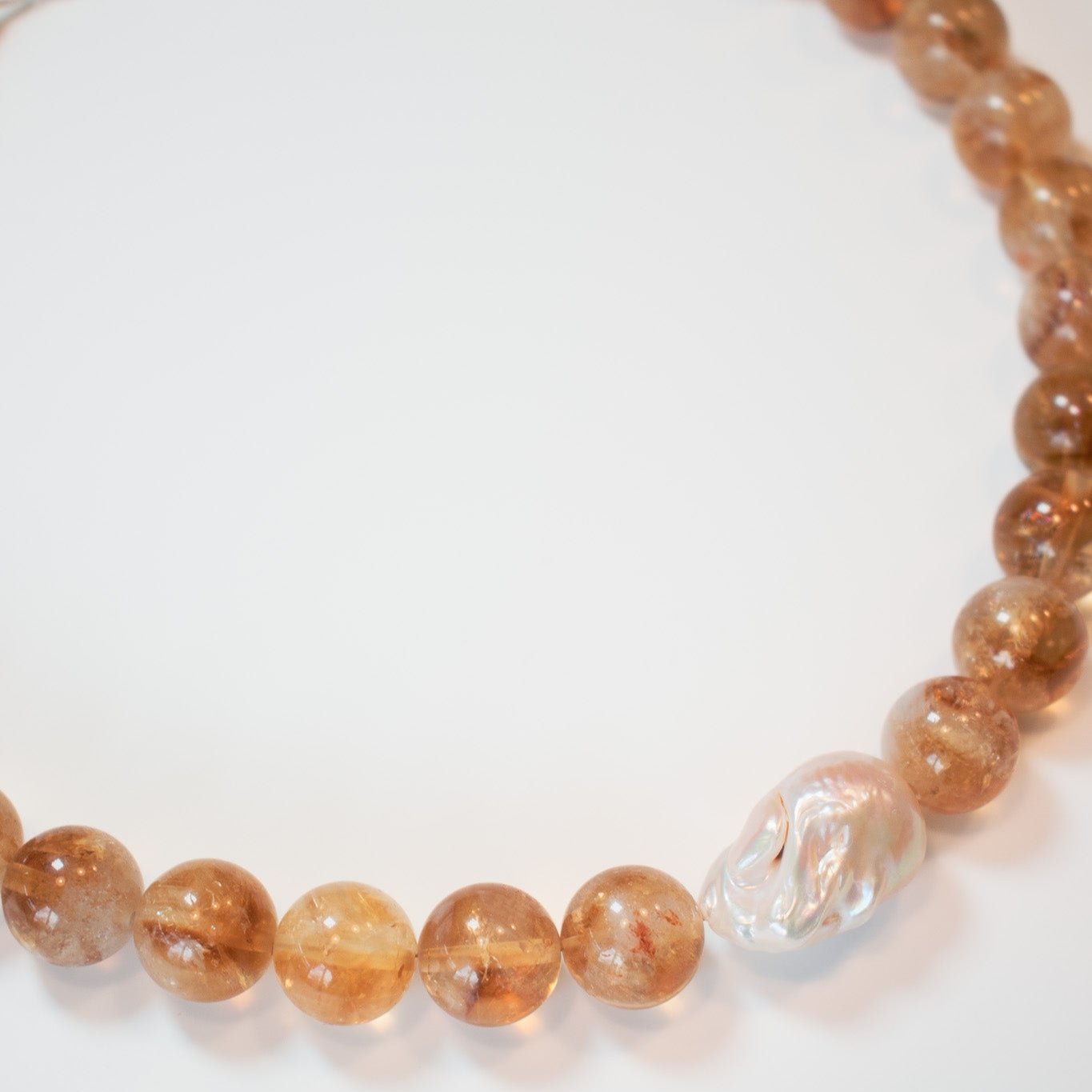 The Pear. Citrine and Pearl Beaded Necklace, showcasing round, translucent brown beads paired with a single baroque pearl, is elegantly displayed in a semi-circle on a white surface.