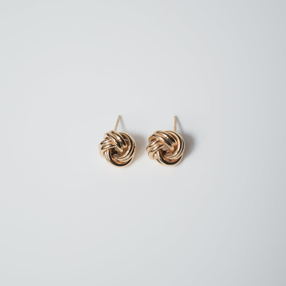 A pair of Pear's Love Knot Earrings in 14K gold glistens on a smooth, light gray background. These statement studs, with their shiny finish, are elegantly arranged side by side.