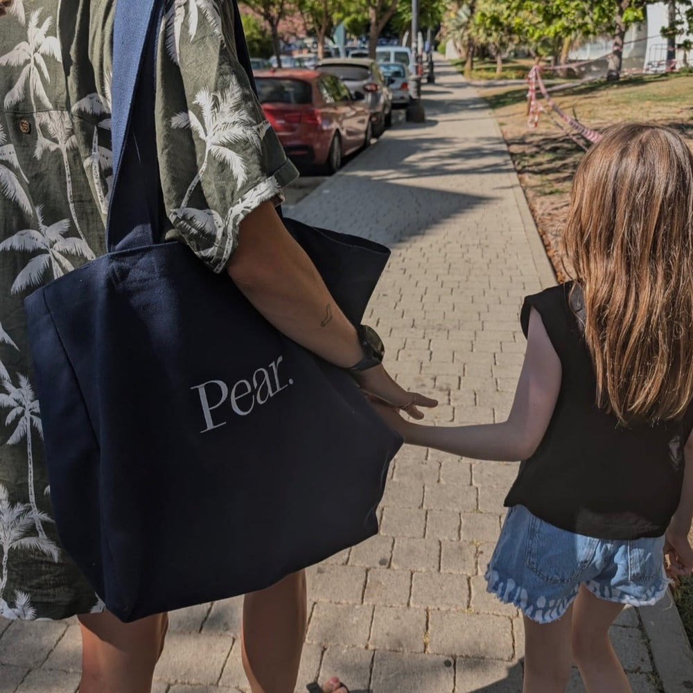 
                      
                        An adult and a child walk hand-in-hand down a sunlit sidewalk. The adult carries an Everyday Tote Bag by Pear. The child wears a black top and denim shorts, while the adult sports a green shirt with a palm tree pattern. Trees and parked cars line the street.
                      
                    