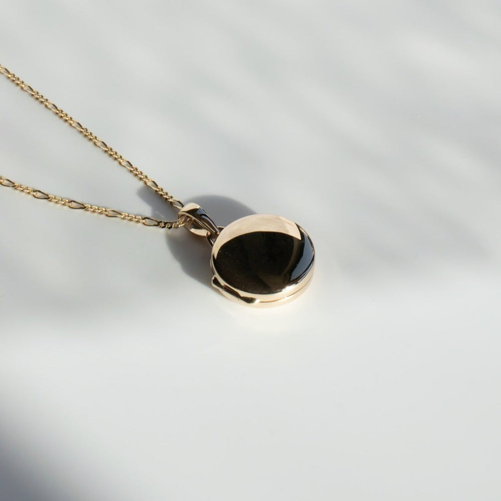 
                      
                        A close-up shot of the Pear Round Locket necklace on a delicate Figaro chain. The pendant is circular, featuring a black gemstone set in the center. Beside the gemstone, there's a 14K gold locket charm. The necklace rests on a white surface with soft shadows falling across it.
                      
                    