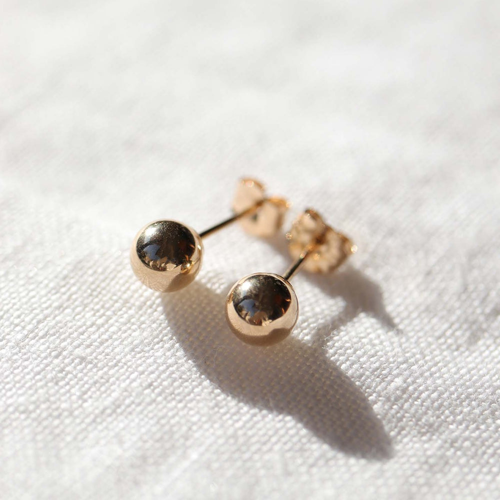 
                      
                        A pair of The Ball Studs by Pear is displayed on a white, textured fabric. Each earring features a polished 14K gold sphere with metallic reflections, attached to a simple post and butterfly clasp. The earrings cast soft shadows on the fabric beneath.
                      
                    