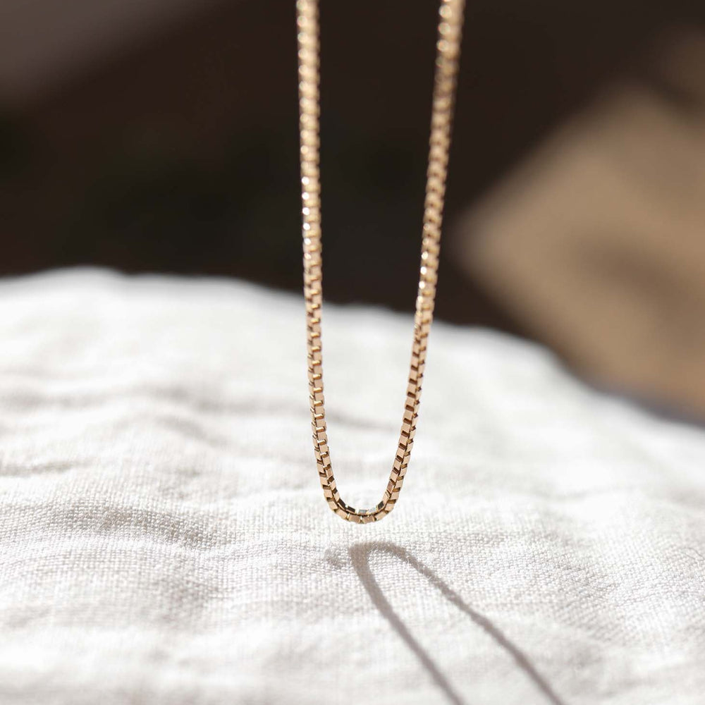 
                      
                        A close-up photograph of The Box Bracelet by Pear, featuring a simple and elegant design. The bracelet, adorned with box-like shapes, is shown hanging and casting a small shadow on a white, textured fabric background. The lighting highlights the bracelet's shiny, polished surface.
                      
                    