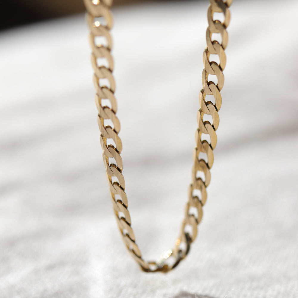 
                      
                        Close-up image of the Pear Solid Curb Bracelet on a white fabric background. The piece features interlocking flat, wide links with a polished finish, giving it a shiny and reflective appearance. The soft light highlights the smooth texture of the gold.
                      
                    