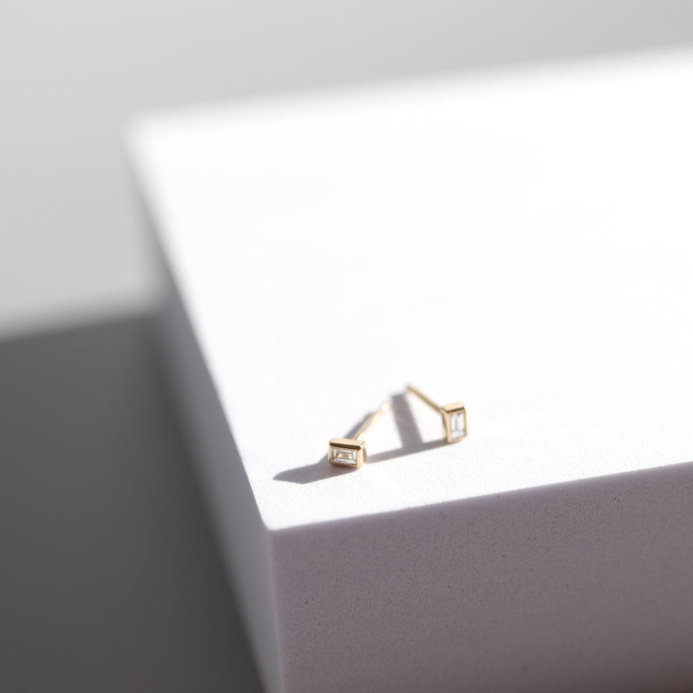 A minimalistic pair of Pear's Diamond Baguette Earrings, each featuring small square decorations, is placed on a white box with clean lines. The shadows and light give a modern, sleek appearance to the setup.