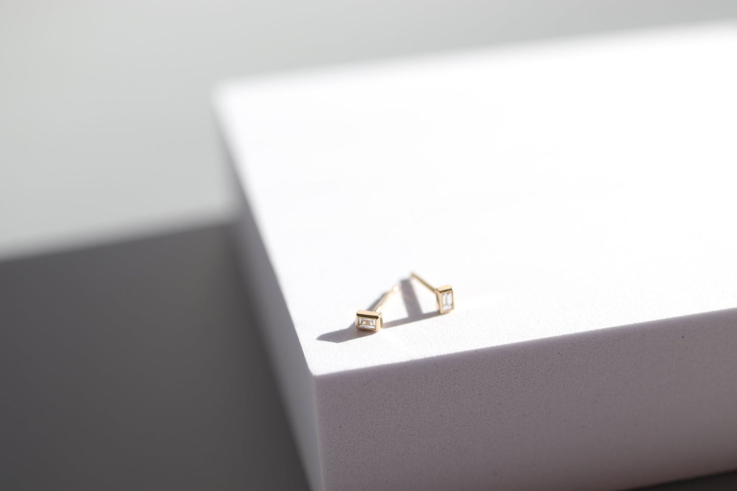 A minimalistic pair of Pear's Diamond Baguette Earrings, each featuring small square decorations, is placed on a white box with clean lines. The shadows and light give a modern, sleek appearance to the setup.