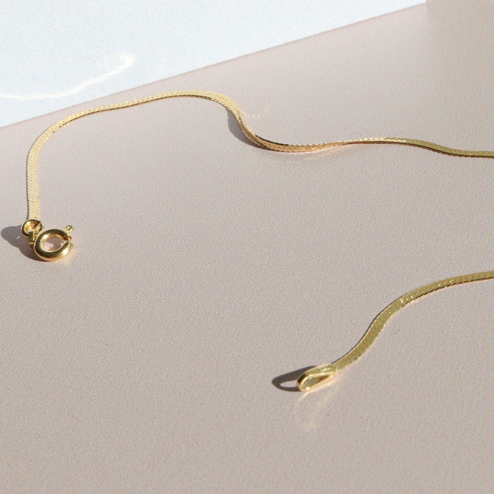 
                      
                        A delicate, thin gold necklace is laid out on a smooth, beige surface. The chain features a small ring clasp and a flat design that reflects light subtly, creating gentle shadows on the surface. Nearby, the Pear. Thin Herringbone Bracelet perfectly complements the elegant display. The background is softly illuminated.


                      
                    