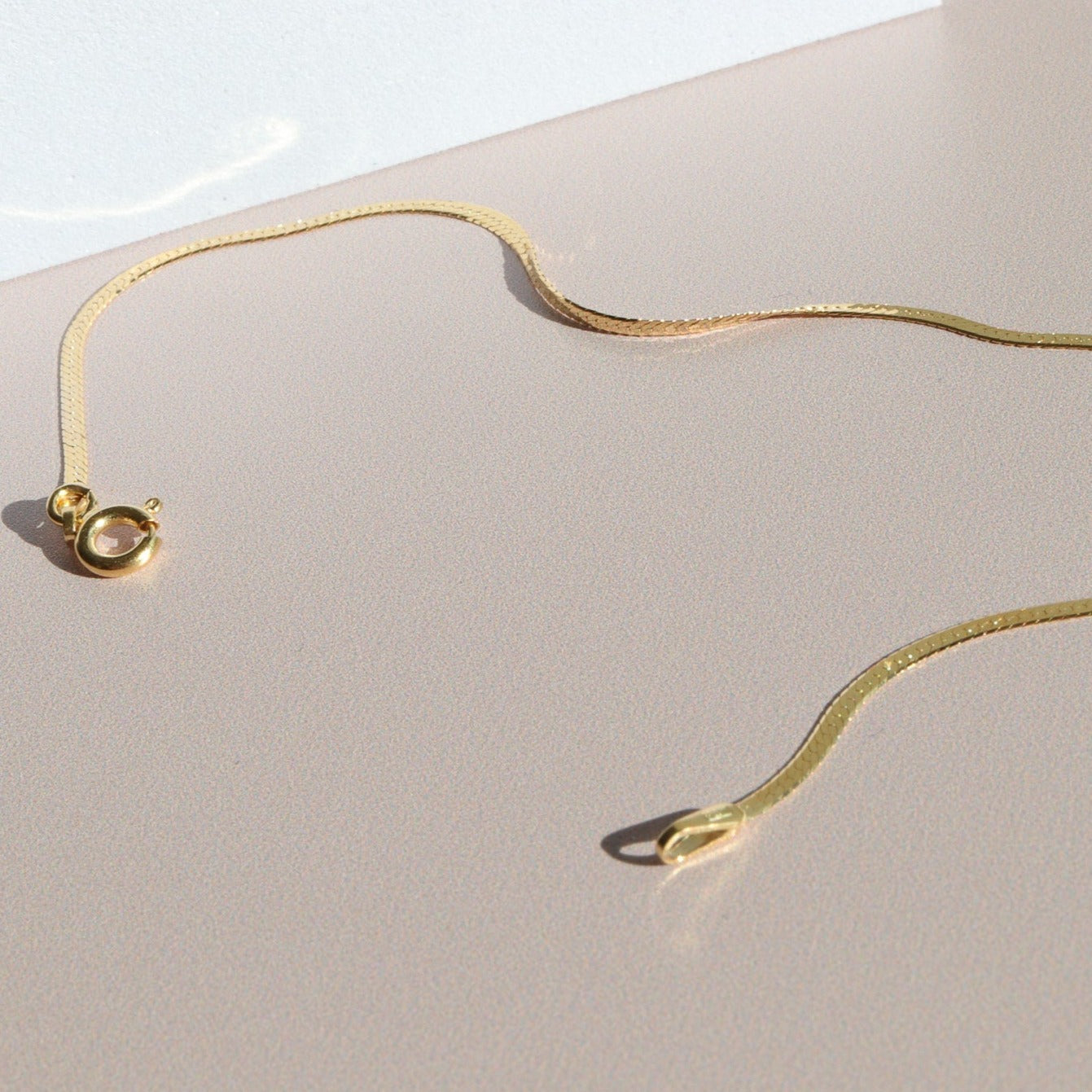 A delicate, thin gold necklace is laid out on a smooth, beige surface. The chain features a small ring clasp and a flat design that reflects light subtly, creating gentle shadows on the surface. Nearby, the Pear. Thin Herringbone Bracelet perfectly complements the elegant display. The background is softly illuminated.

