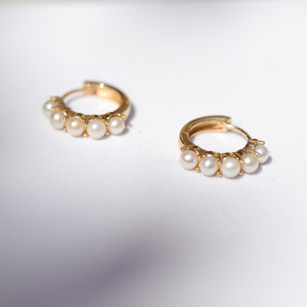 
                      
                        A pair of Pear's Pearl Huggies, crafted from 14K Gold and adorned with small freshwater pearls, are placed on a white surface. The earrings are positioned with the clasps open, showcasing their intricate design and elegant appearance. The lighting highlights the luster of the pearls and the shine of the gold metal.
                      
                    