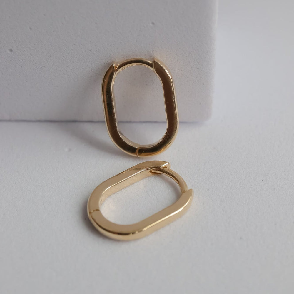 
                      
                        The Pear "Elliot Hoops" are a pair of solid gold earrings with an elongated oval shape, showcased with one earring standing upright against a white background and the other lying flat beside it. Their sleek, minimalist design is complemented by a practical latch-style hinge.
                      
                    