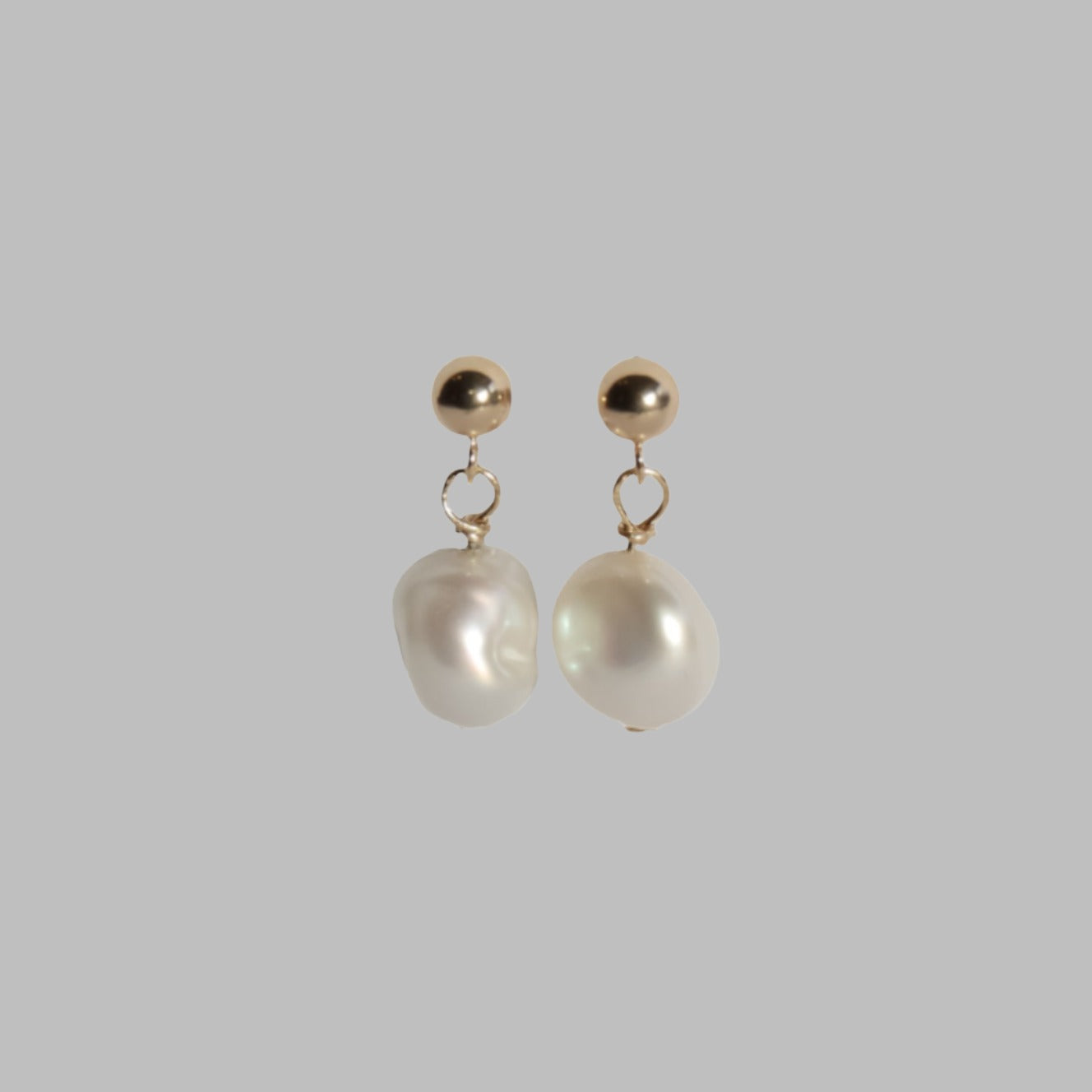 Introducing the Pearl Boule Earrings by Pear: a pair of elegant earrings showcasing round 14K gold balls at the top, from which white, irregularly shaped freshwater pearls gracefully dangle. The solid light gray background enhances the simple yet sophisticated design of these exquisite earrings.