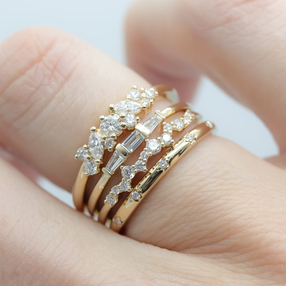 Close-up of a hand wearing the elegant Edith Diamond Ring by Pear, featuring four gold bands adorned with various diamonds. The rings have different designs, including round diamonds, three-diamond baguette arrangements, and marquise-shaped diamonds set in intricate patterns. The stacked rings on one finger showcase their refined beauty.