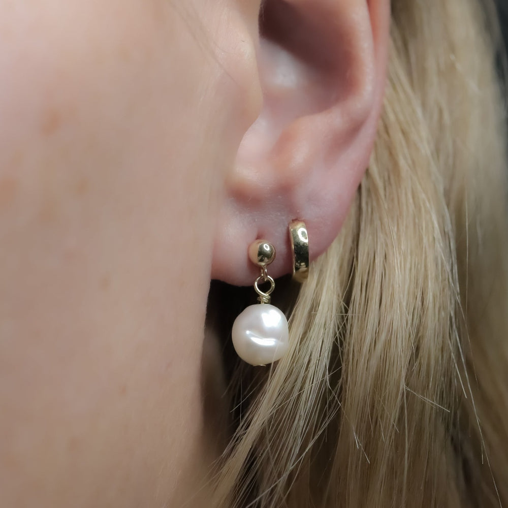 
                      
                        Close-up of a person's ear wearing two earrings. The first is a small gold hoop adorned with 14K gold balls, and the second is a slightly larger hoop featuring a freshwater pearl drop. The ear is partially visible against a blurred background, highlighting the elegant Pearl Boule Earrings by Pear.
                      
                    