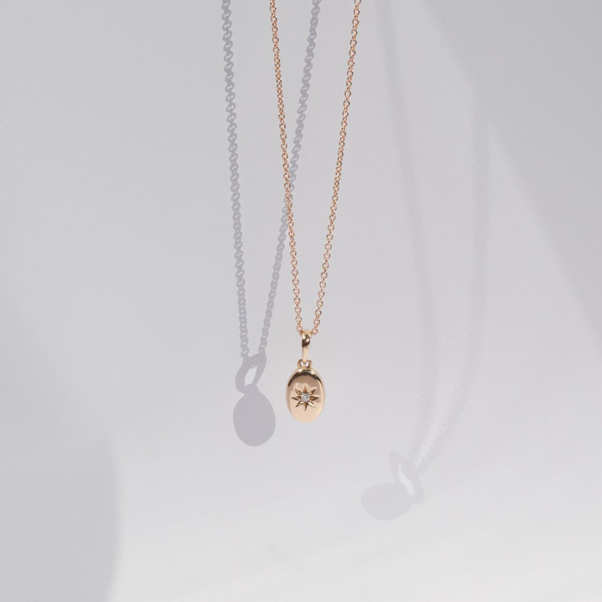 A delicate gold necklace from Pear's Astrid Necklace collection hangs gracefully against a white background. The 14k gold pendant, oval in shape, is accented with a diamond star design at its center and casts a soft shadow. The overall aesthetic is minimalistic and elegant.