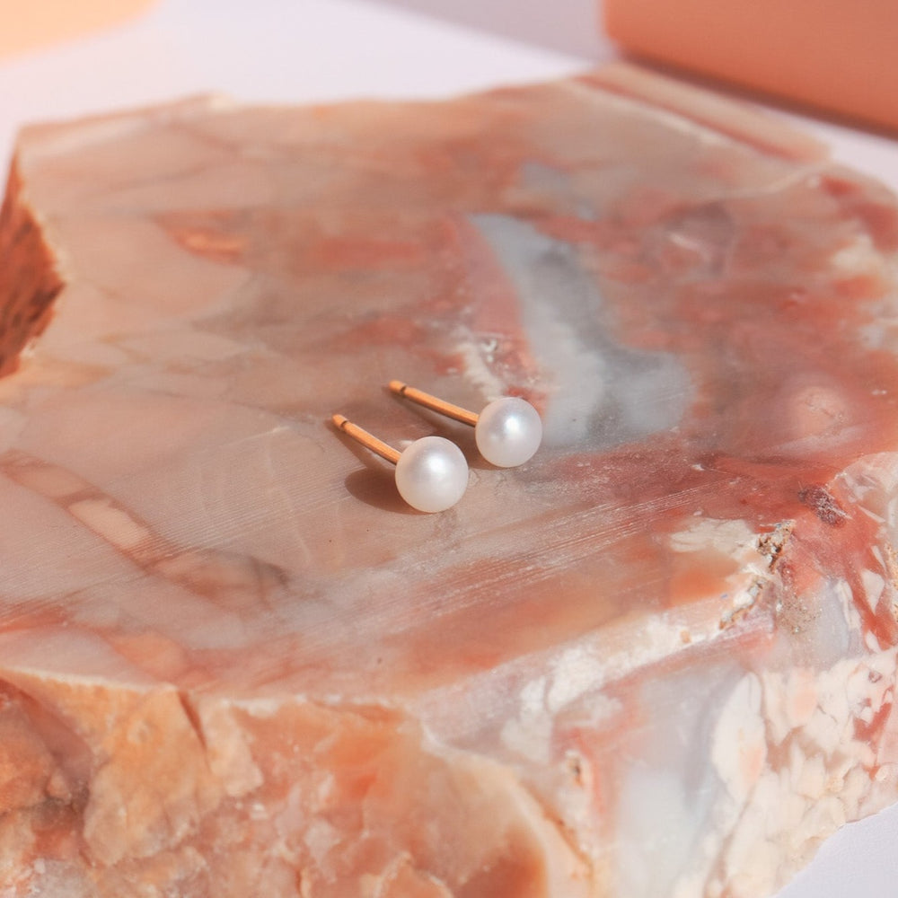 
                      
                        A pair of Pear Pearl Studs, featuring delicate freshwater pearls and 14K gold posts, are elegantly set on a polished, multicolored stone surface with pink, beige, and white hues. Soft light accentuates the pearls' luster and the intricate patterns of the stone.
                      
                    