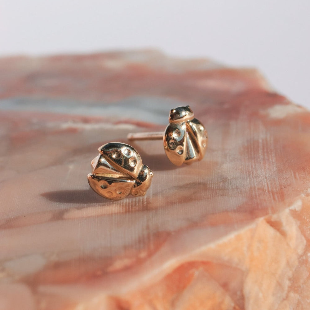 
                      
                        A pair of Pear's Ladybug Studs, crafted from solid 14K gold, rest on a pink and beige marble surface. These polished earrings feature intricate details that capture the texture and spots typical of a ladybug's back. The marble background provides a soft, elegant contrast to the jewelry.
                      
                    