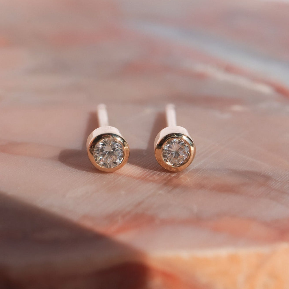 
                      
                        Close-up of a pair of Pear's Mini Bezel Diamond Studs in 14K Solid Gold with round, transparent gemstones, set against a soft pink marble background. The earrings have a simple and elegant design, featuring a bezel setting that secures the natural diamonds.
                      
                    