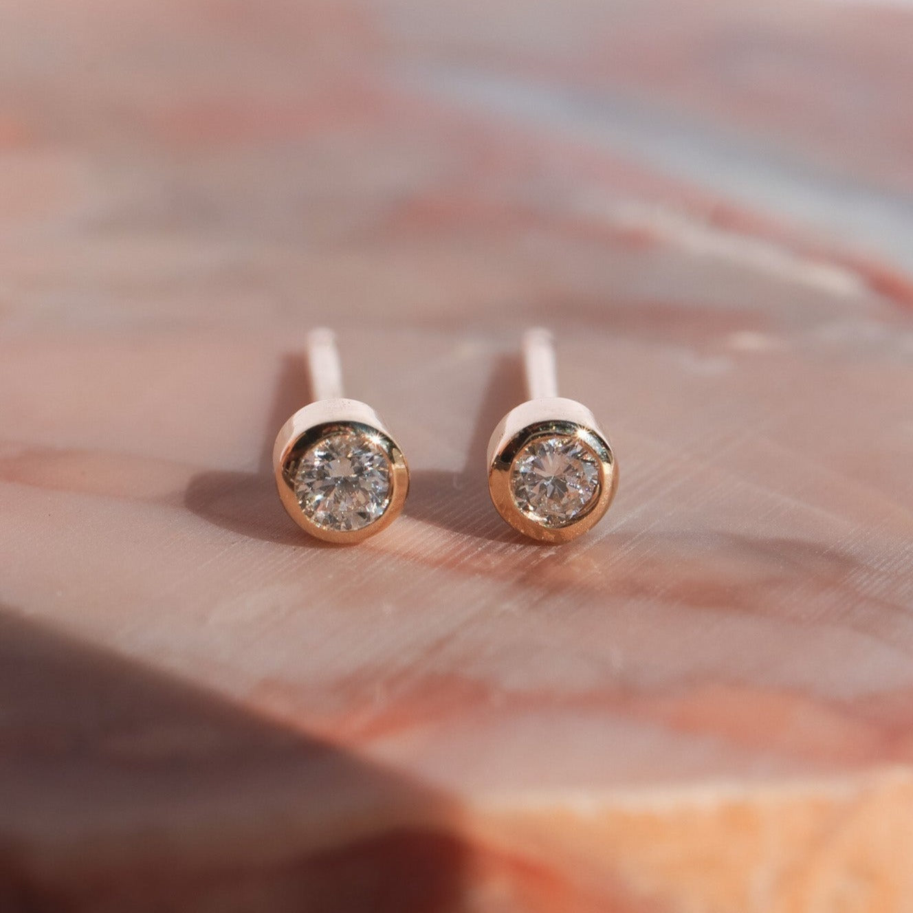 Close-up of a pair of Pear's Mini Bezel Diamond Studs in 14K Solid Gold with round, transparent gemstones, set against a soft pink marble background. The earrings have a simple and elegant design, featuring a bezel setting that secures the natural diamonds.