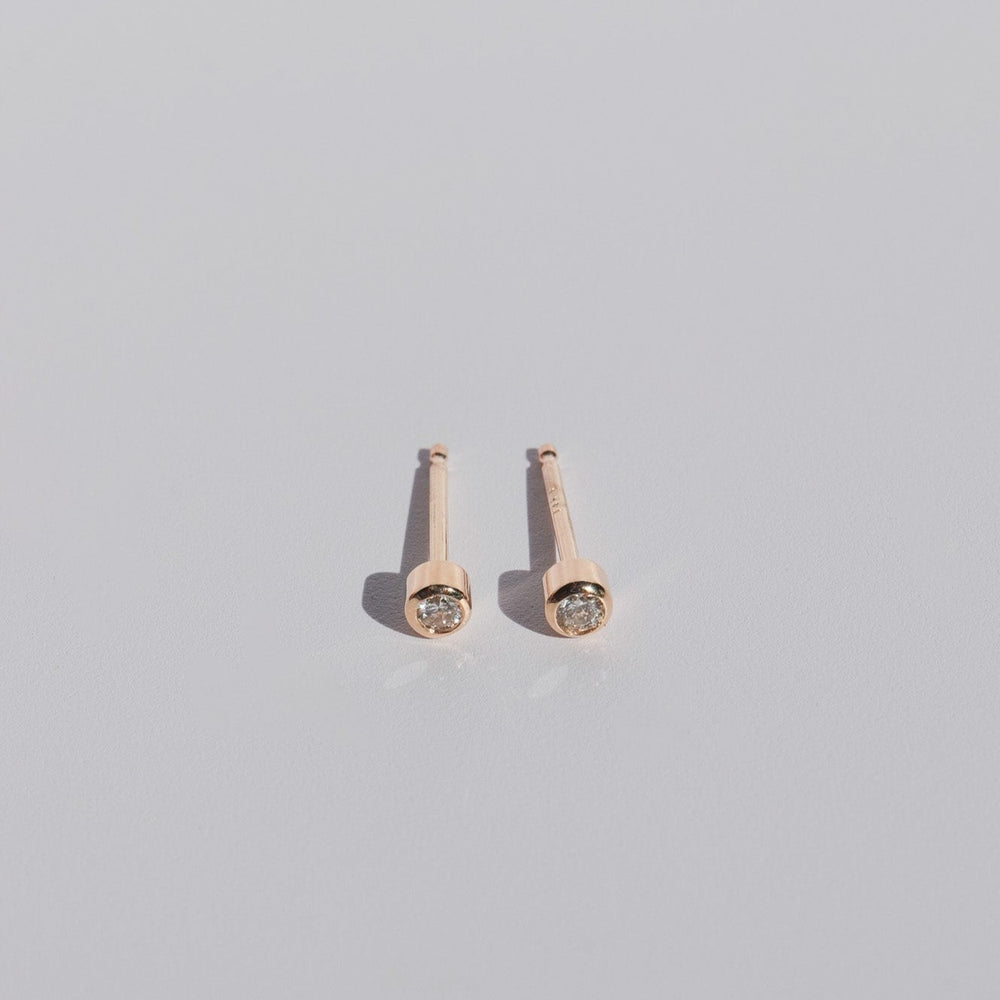 A pair of Mini Bezel Diamond Studs by Pear in 14K Solid Gold featuring round, clear natural diamonds on a plain, light gray background. The earrings are positioned parallel to each other, highlighting their shiny and minimalistic design.