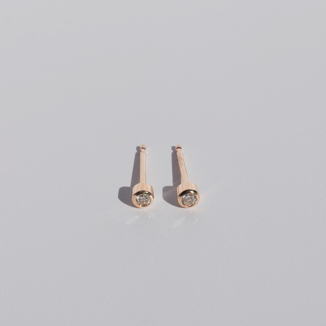 A pair of Mini Bezel Diamond Studs by Pear in 14K Solid Gold featuring round, clear natural diamonds on a plain, light gray background. The earrings are positioned parallel to each other, highlighting their shiny and minimalistic design.