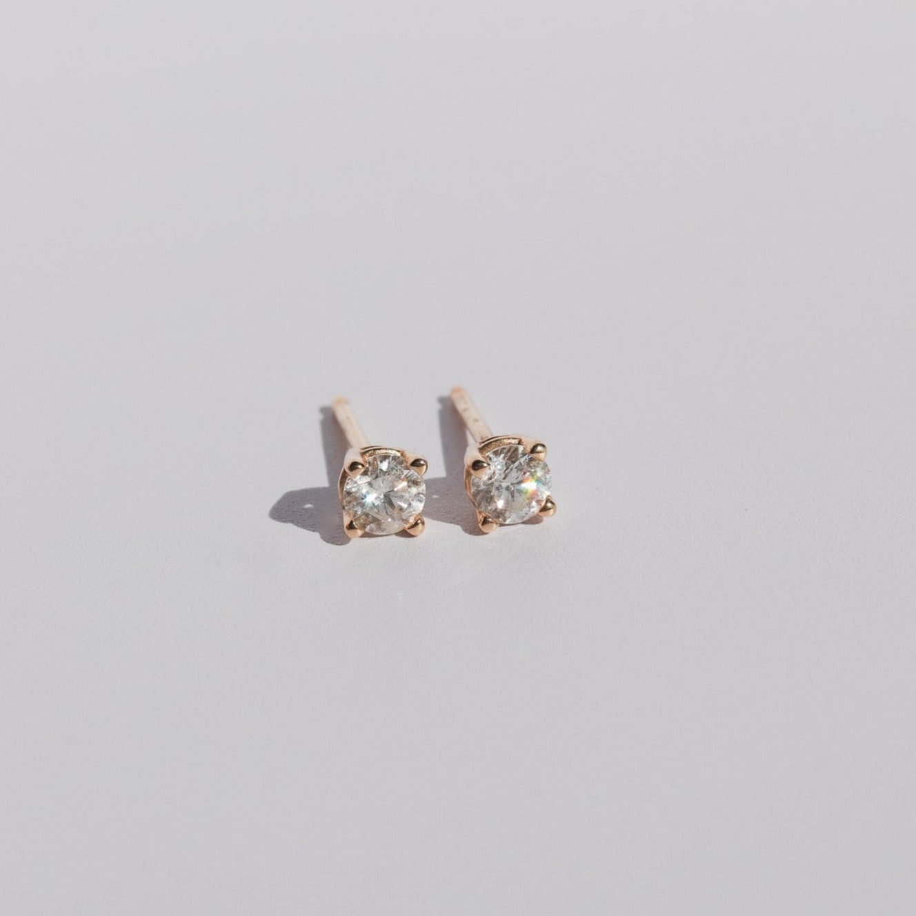 The Diamond Prong Studs by Pear feature round, natural brilliant cut diamonds set in gold prongs. Positioned side by side on a plain, light-colored surface, the earrings highlight their minimalist design and diamonds of SI1 grading quality.