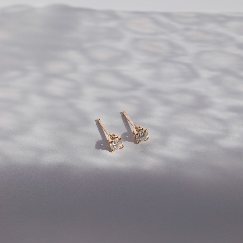 
                      
                        A pair of Pear's Diamond Prong Studs, featuring natural brilliant cut diamonds set in clear gemstone settings, is placed on a smooth white surface. Shadows with a subtle pattern play around the diamond earrings, creating a visually textured background.
                      
                    