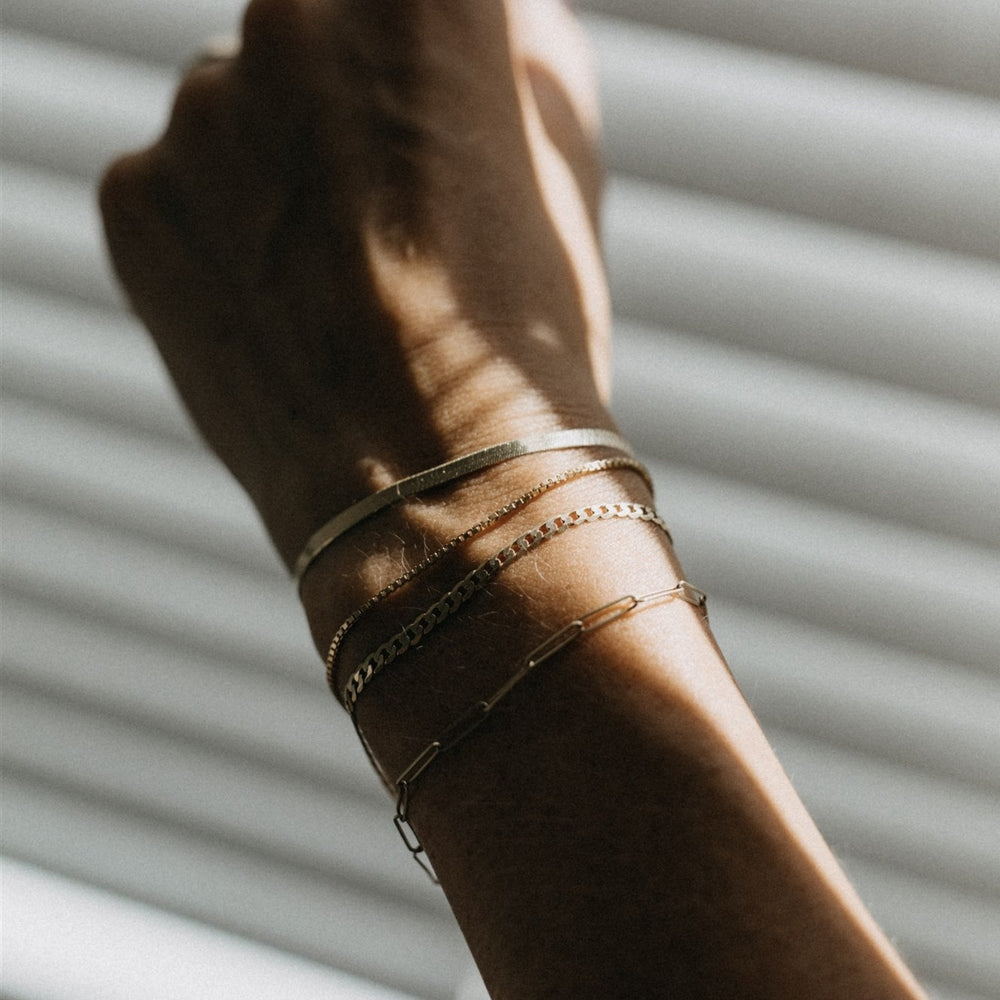 
                      
                        A close-up of a person's wrist adorned with three delicate gold bracelets, including The Box Bracelet by Pear, featuring a Venetian chain with a Spring Ring Clasp. The hand is slightly clenched and raised, with light streaming through horizontal blinds in the background, casting box-like shadows across the scene.
                      
                    