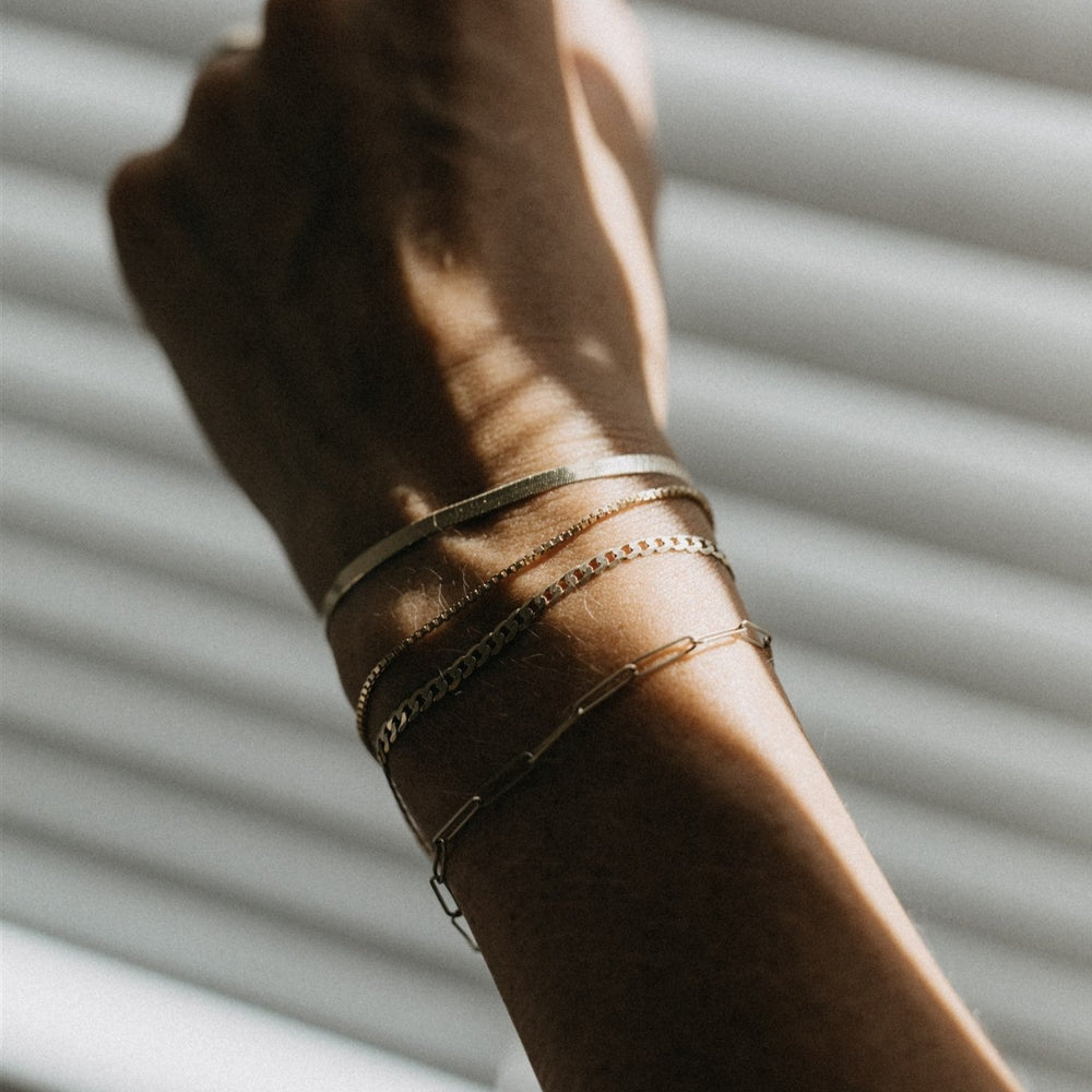 
                      
                        A close-up shot of a forearm with sunlight and shadows from window blinds cast onto the skin. The arm is adorned with three slim Pear Solid Curb Bracelets, showcasing varying chain styles including one with interlocking links. The background is softly lit, creating horizontal striped shadows.
                      
                    