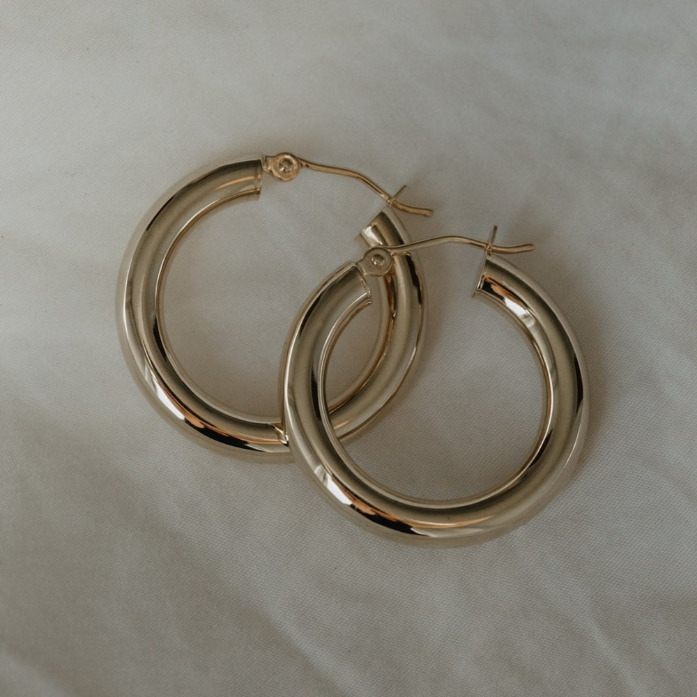 
                      
                        A pair of Pear.'s Thick Tube Hoops, made from lightweight, solid 14K gold, is displayed on a light, textured fabric background. The thick hoops have a smooth, shiny surface and are secured with hook clasps.
                      
                    