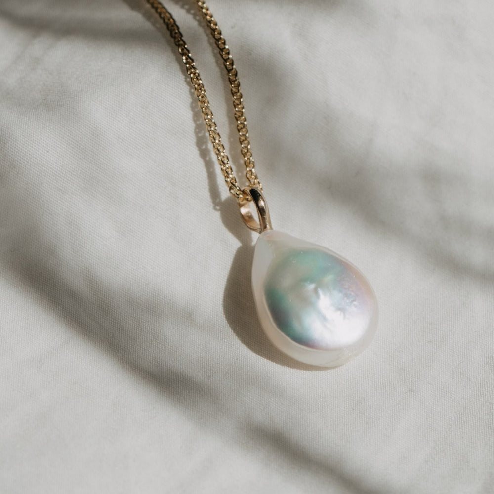 
                      
                        A close-up of the Baroque Pearl Necklace by Pear, featuring a 14K yellow gold chain with a large, teardrop-shaped pearlescent pendant. The necklace is set against a light, textured fabric background with soft shadows.
                      
                    