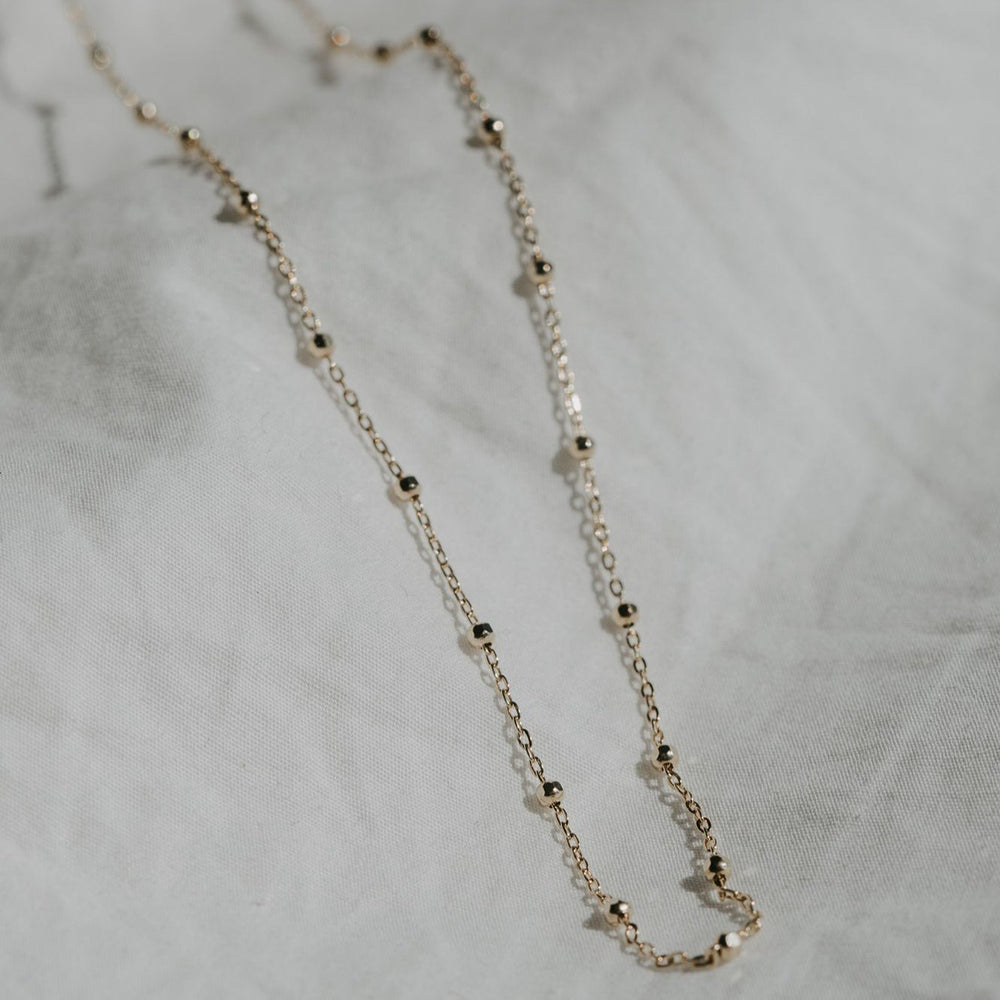 
                      
                        The Beaded Chain by Pear is laid out flat on a light, textured fabric background, showcasing its delicate gold beaded cable chain. Small, evenly spaced faceted beads adorn the necklace, and the soft sunlight casts gentle shadows that highlight the elegance and simplicity of this exquisite piece, secured by a lobster clasp.
                      
                    