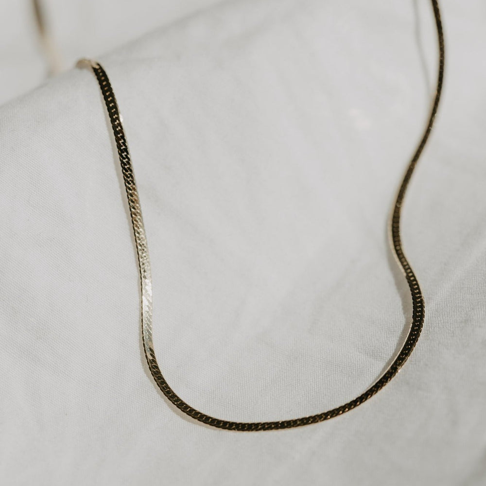 A thin, elegant herringbone bracelet from Pear., adorned with a subtle textured pattern, is displayed against a soft, white fabric background. Showcasing a sleek style, the bracelet is gently curved, highlighting its delicate nature and simple yet sophisticated design.